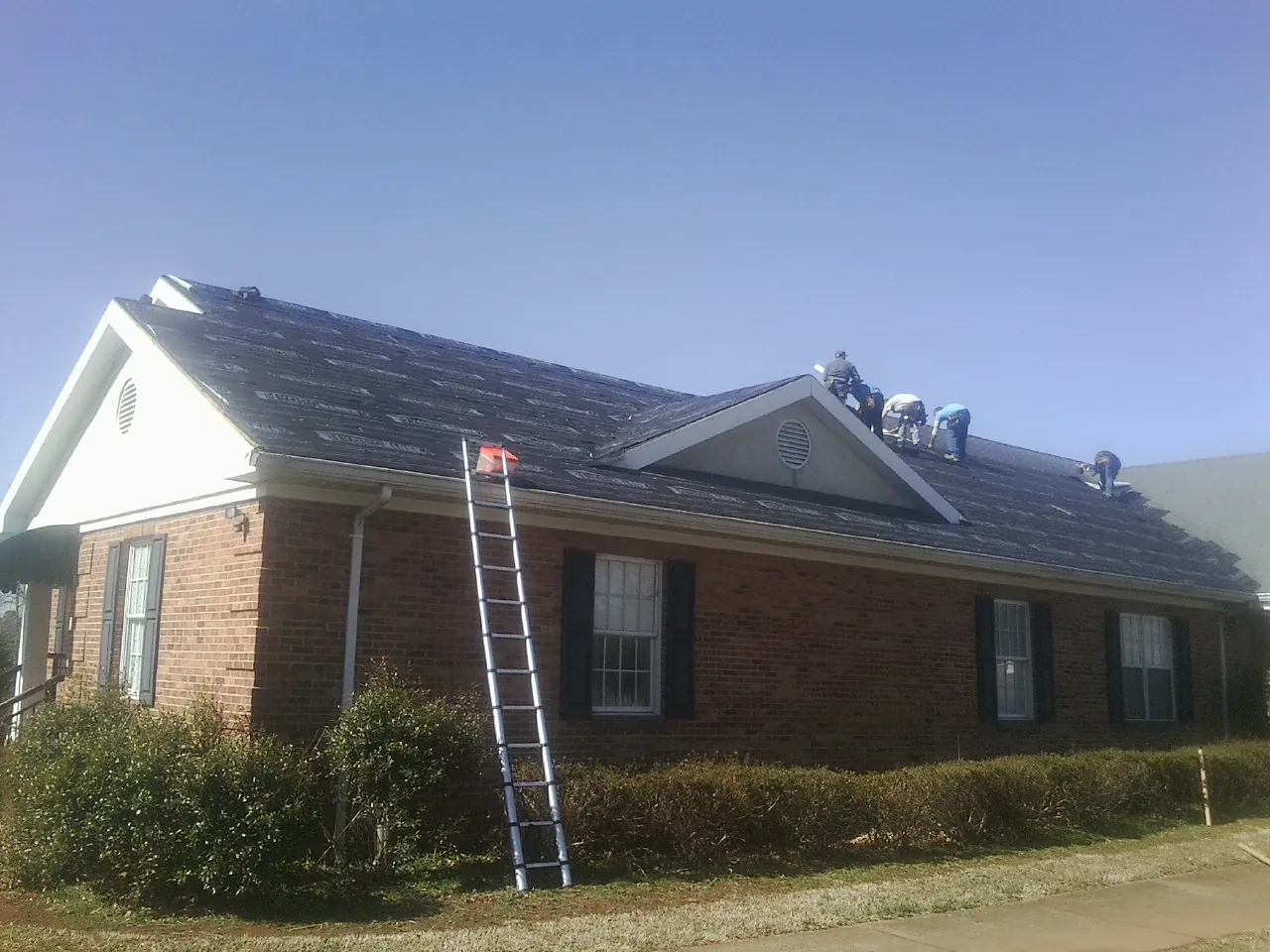 Crew tearing shingles off a brick ranch roof with a ladder set — Emergency Roof Repair work by our crew in Pineland