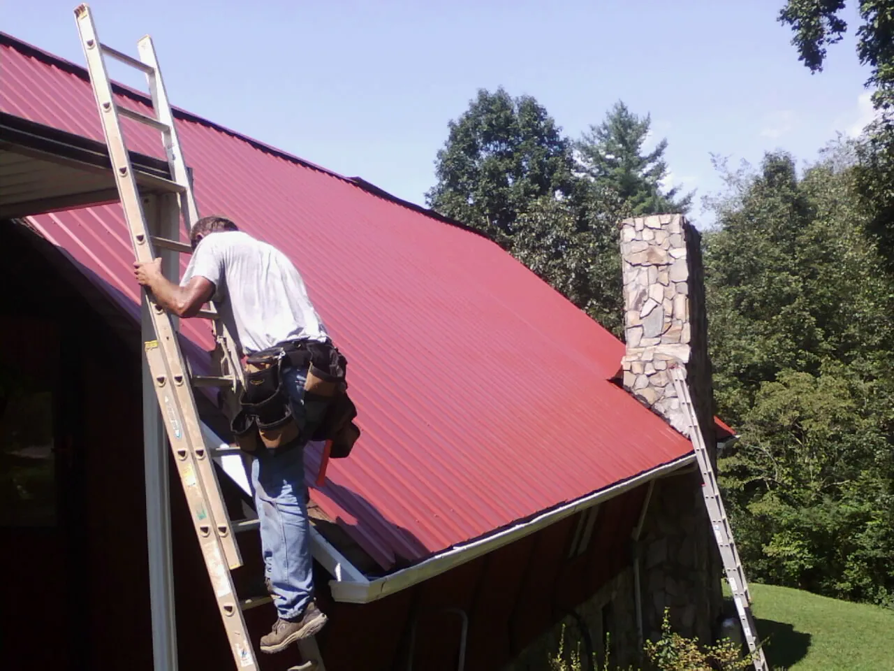 Roofer on a ladder beside a red metal roof and stone chimney — Soffit & Fascia Repair by Roofing Brothers in Pineland